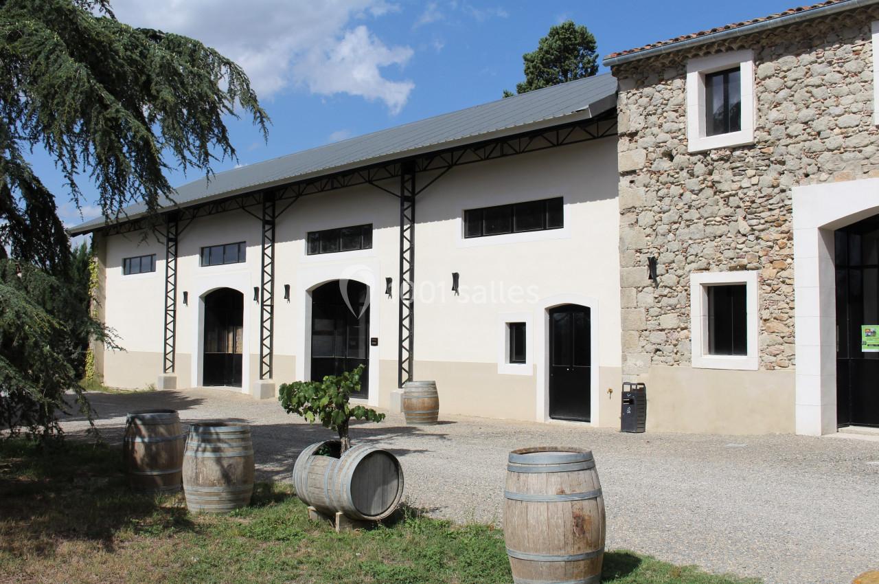 FACADE CORUM Bâtiment en pierre et crépi avec des portes noires, entouré de barriques en bois et d'arbres sous un ciel dégagé.