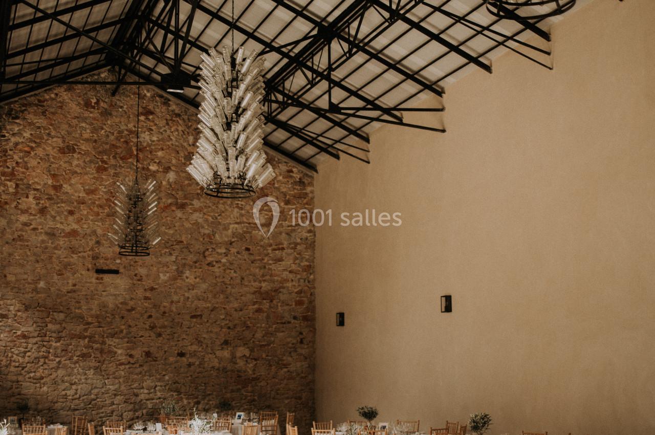 LUSTRES DE NOTRE SALLE CORUM Salle de réception lumineuse avec tables rondes dressées, chaises en bois clair et lustres suspendus sous une verrière.