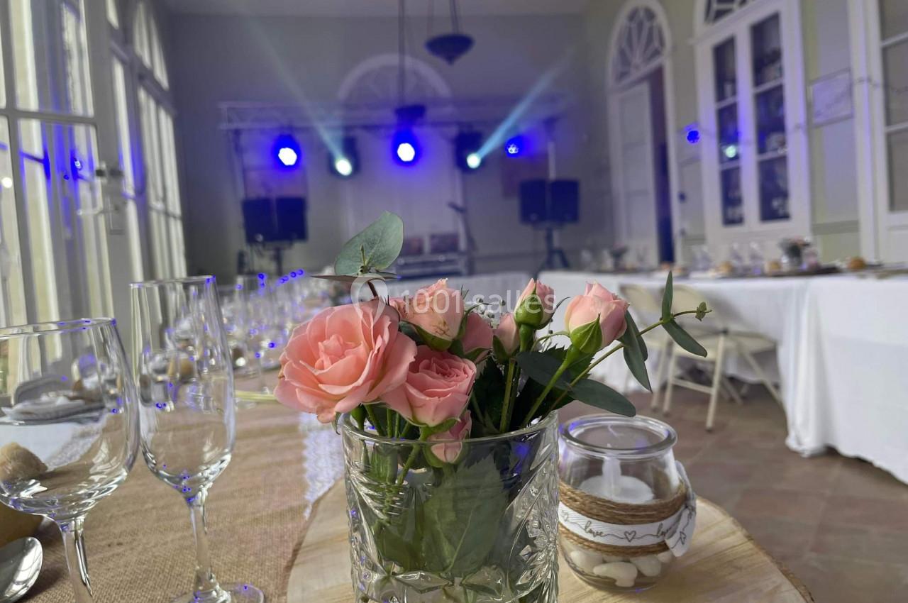 MARIAGE DANS LE PERISTYLE DU CHATEAU Vase de roses roses sur une table décorée, avec une salle lumineuse et une scène éclairée en arrière-plan.