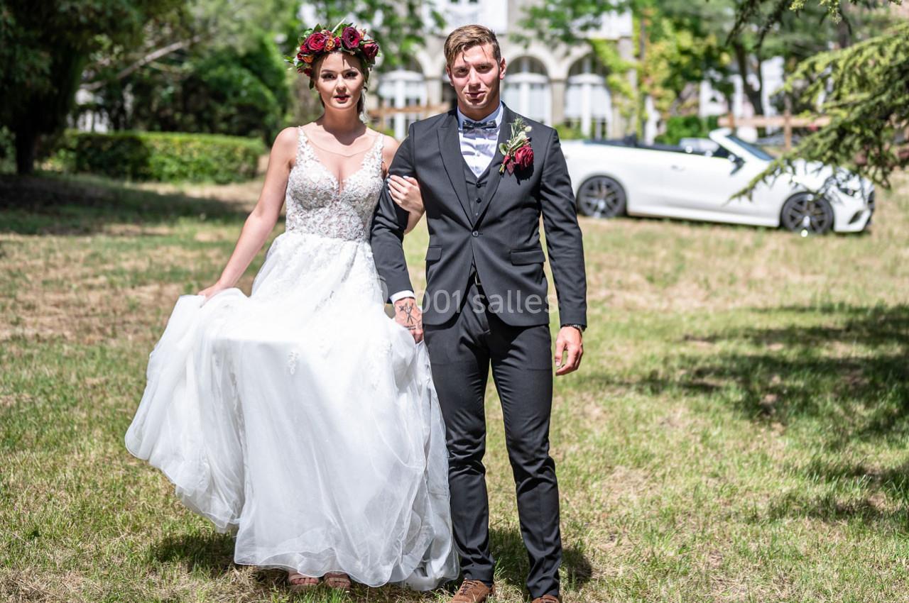 PARC DU CHATEAU Un couple en tenue de mariage marche sur une pelouse, avec une voiture blanche décapotable en arrière-plan.