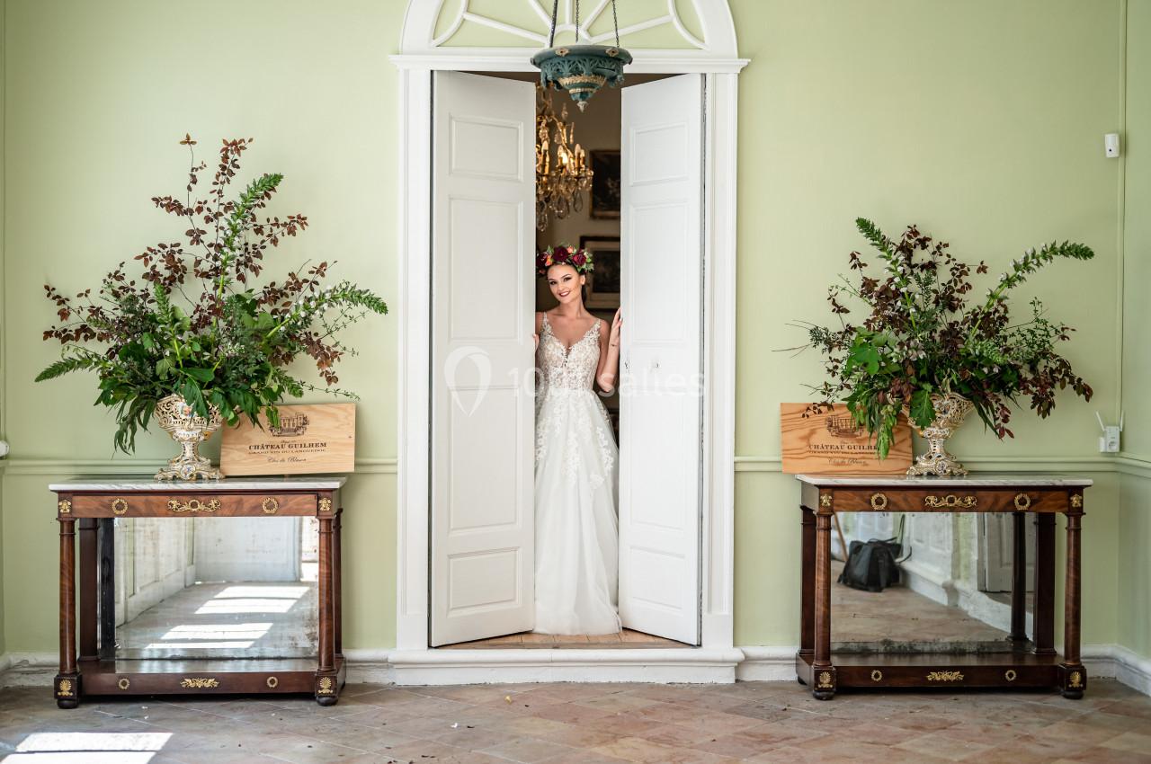 PERISTYLE DU CHATEAU Une mariée en robe blanche se tient dans l'encadrement d'une porte blanche, entourée de décorations florales.