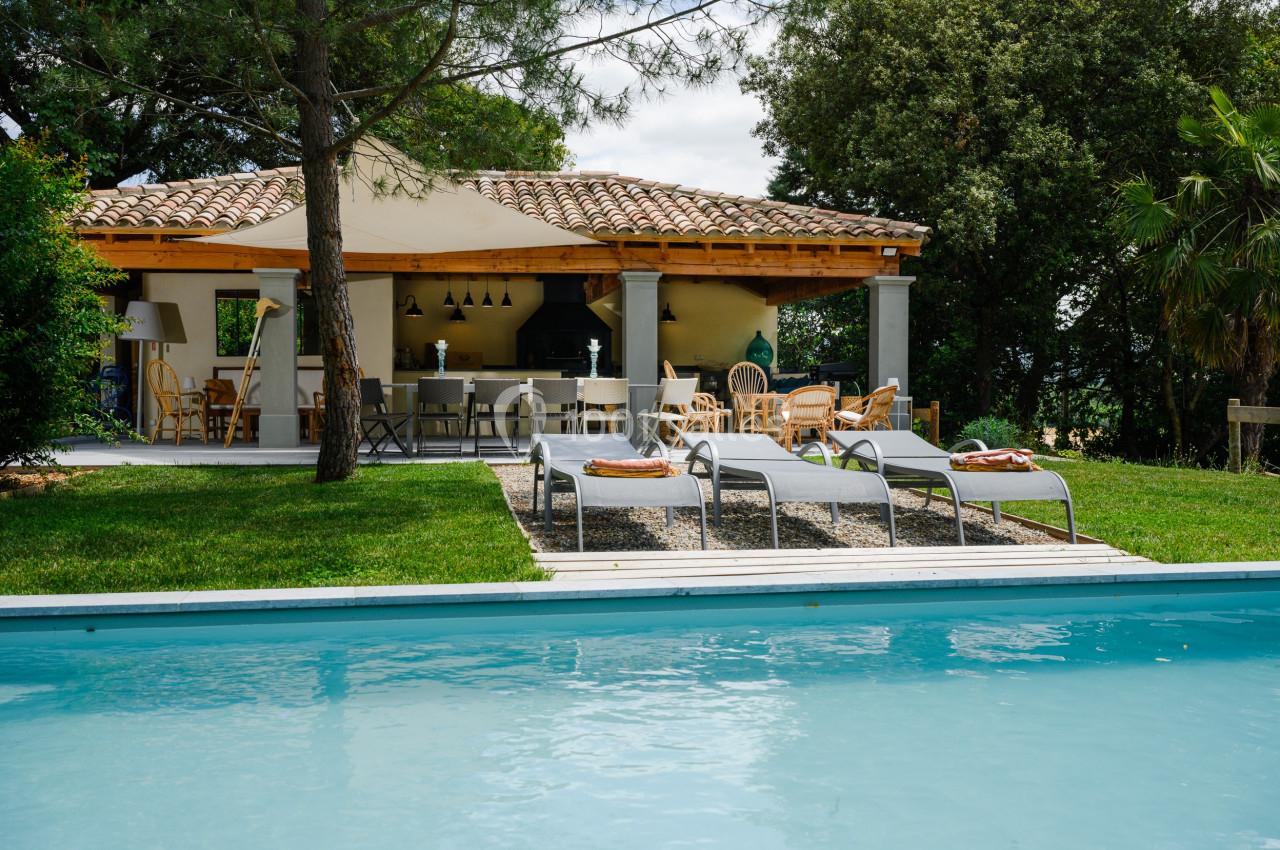 POOL HOUSE DE 100 M2 Terrasse avec mobilier en bois sous une pergola, entourée de verdure, près d'une piscine et de chaises longues.