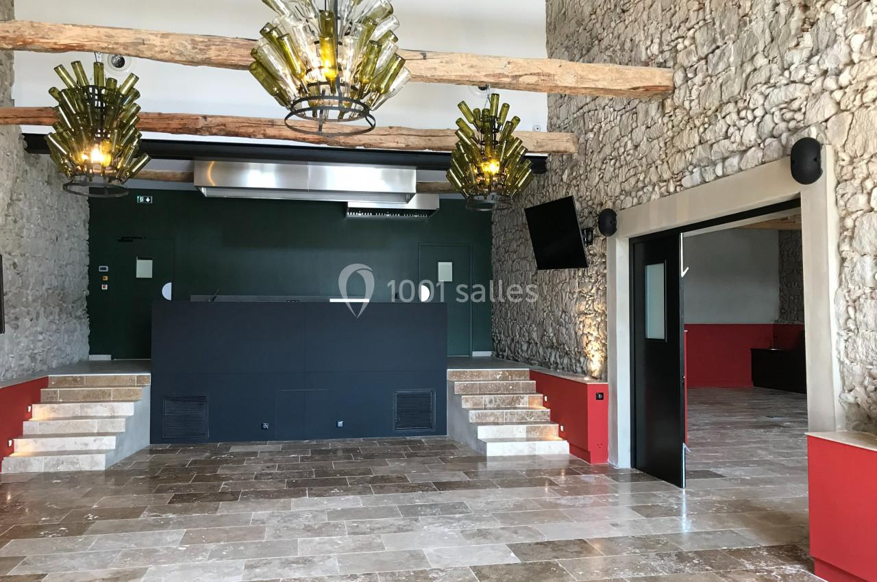 SALLE COMEDIS Salle intérieure avec murs en pierre, lustres en bouteilles recyclées, sol en pierre et comptoir noir central.