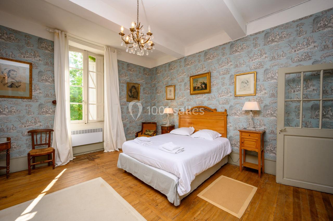 Chambre lumineuse avec lit double, murs tapissés de motifs, parquet en bois, et fenêtre donnant sur un espace verdoyant.