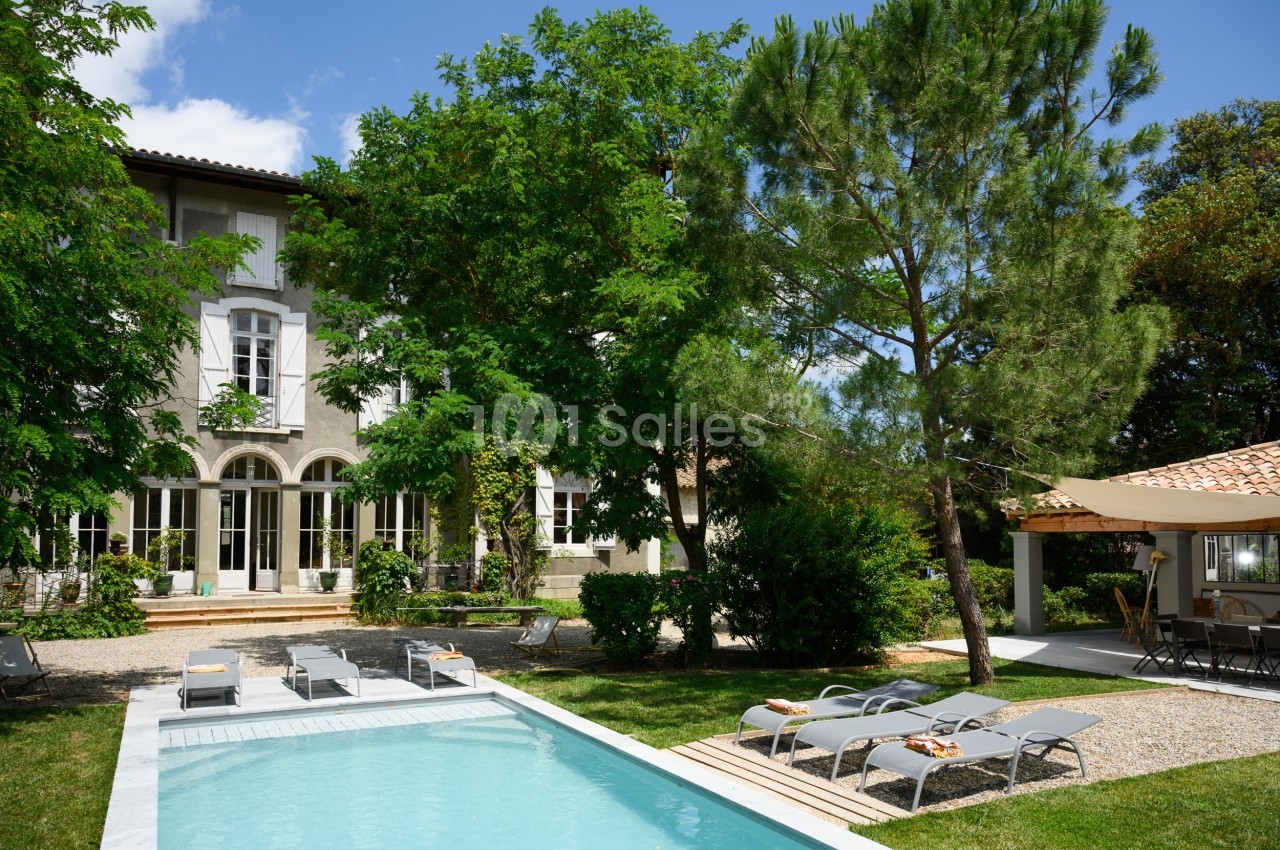Maison avec façade claire, jardin arboré, piscine entourée de transats et terrasse ombragée sous un ciel dégagé.