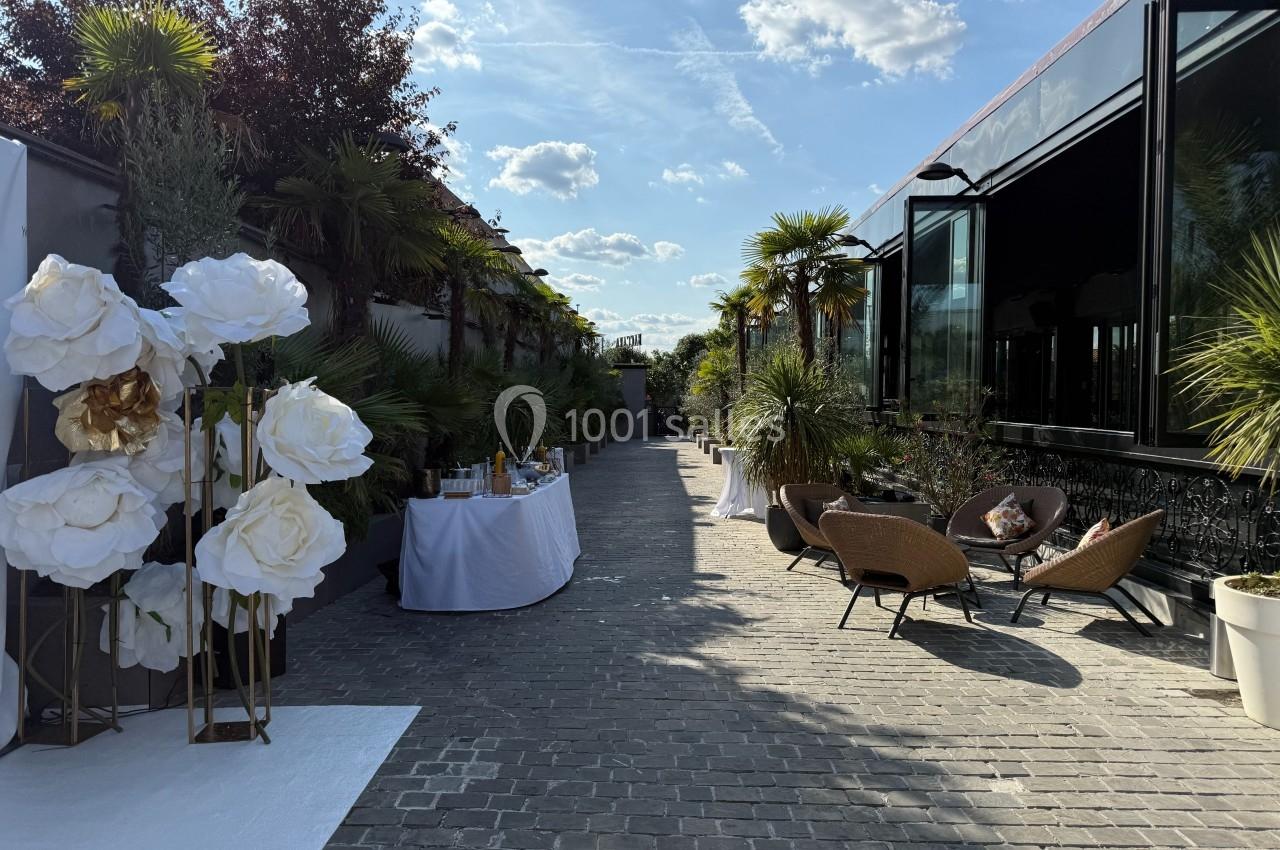 Allée pavée bordée de plantes, avec tables décorées et fauteuils en osier, sous un ciel ensoleillé.