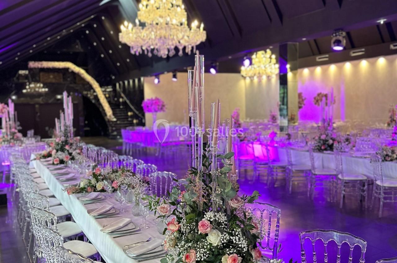 Salle de réception élégante avec tables décorées de fleurs roses et chandeliers lumineux suspendus.