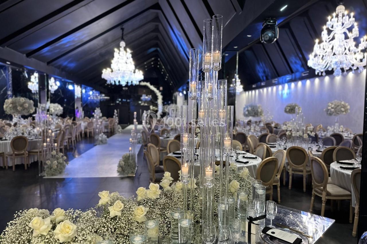 Salle de réception élégante avec tables décorées, chandeliers suspendus et bougies sur des supports en verre.