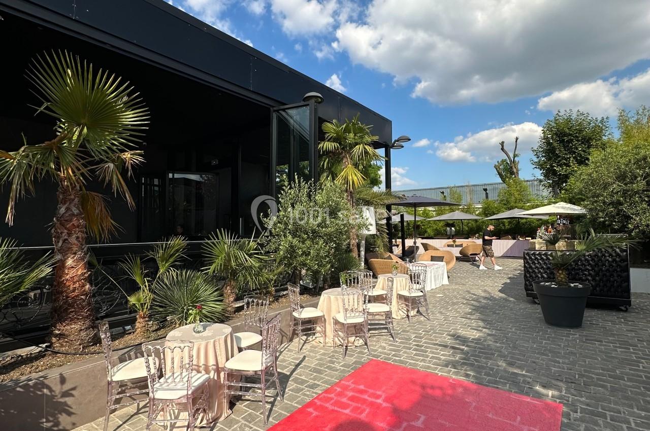 Terrasse extérieure avec tables et chaises, entourée de plantes et palmiers, sous un ciel partiellement nuageux.