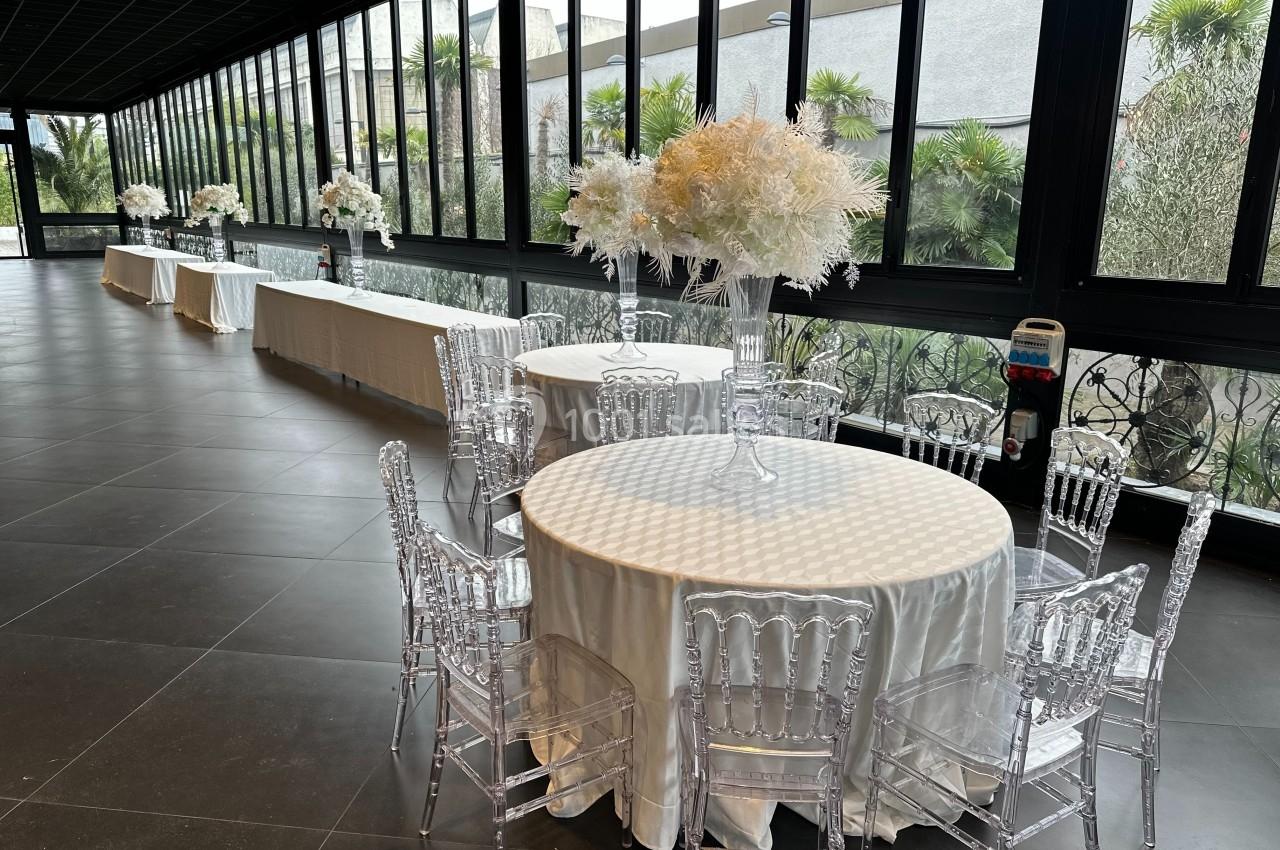 Salle lumineuse avec grandes baies vitrées, tables rondes dressées avec nappes blanches et chaises transparentes.