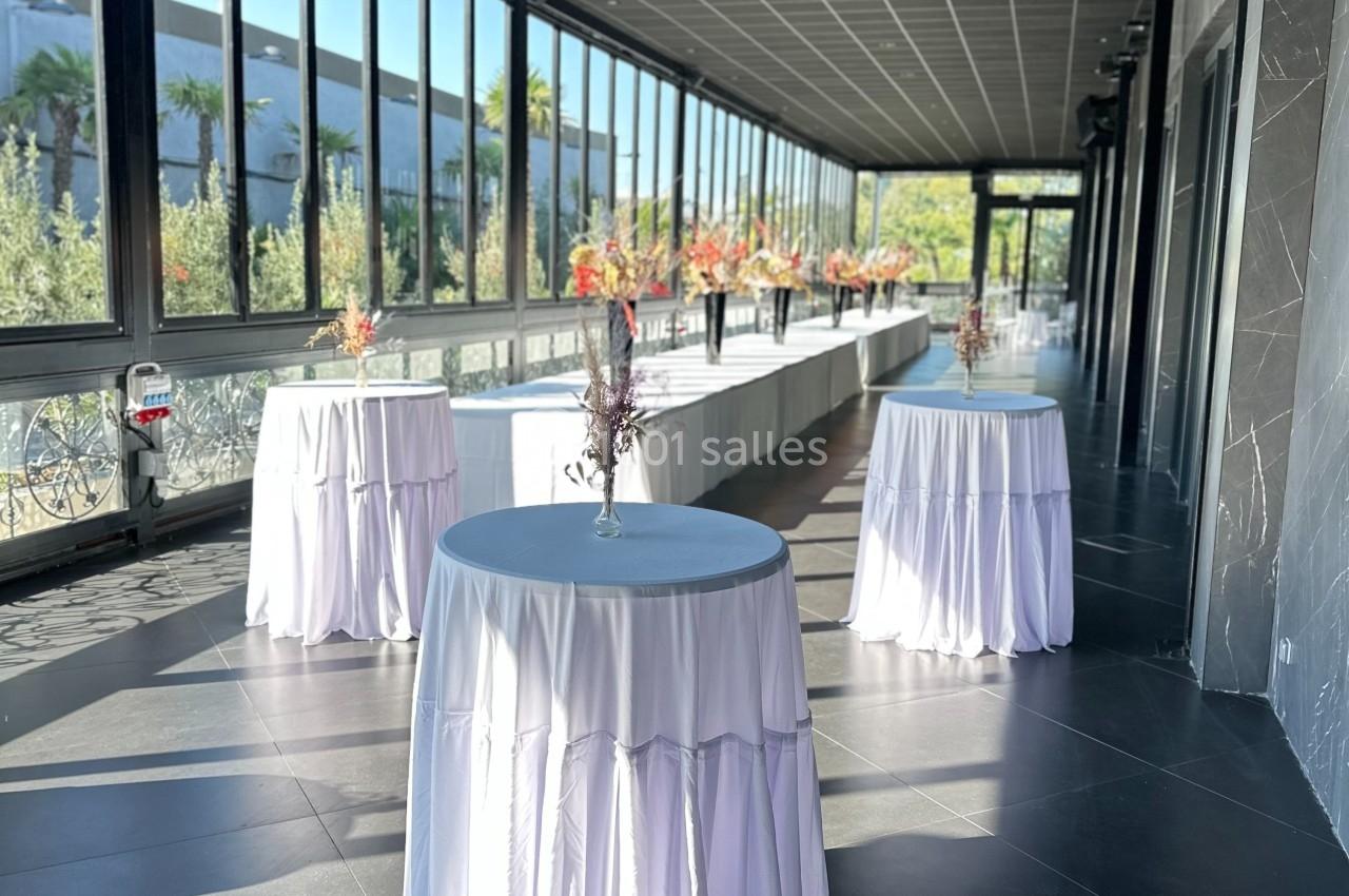 Salle lumineuse avec grandes baies vitrées, tables hautes drapées de blanc et décorations florales.