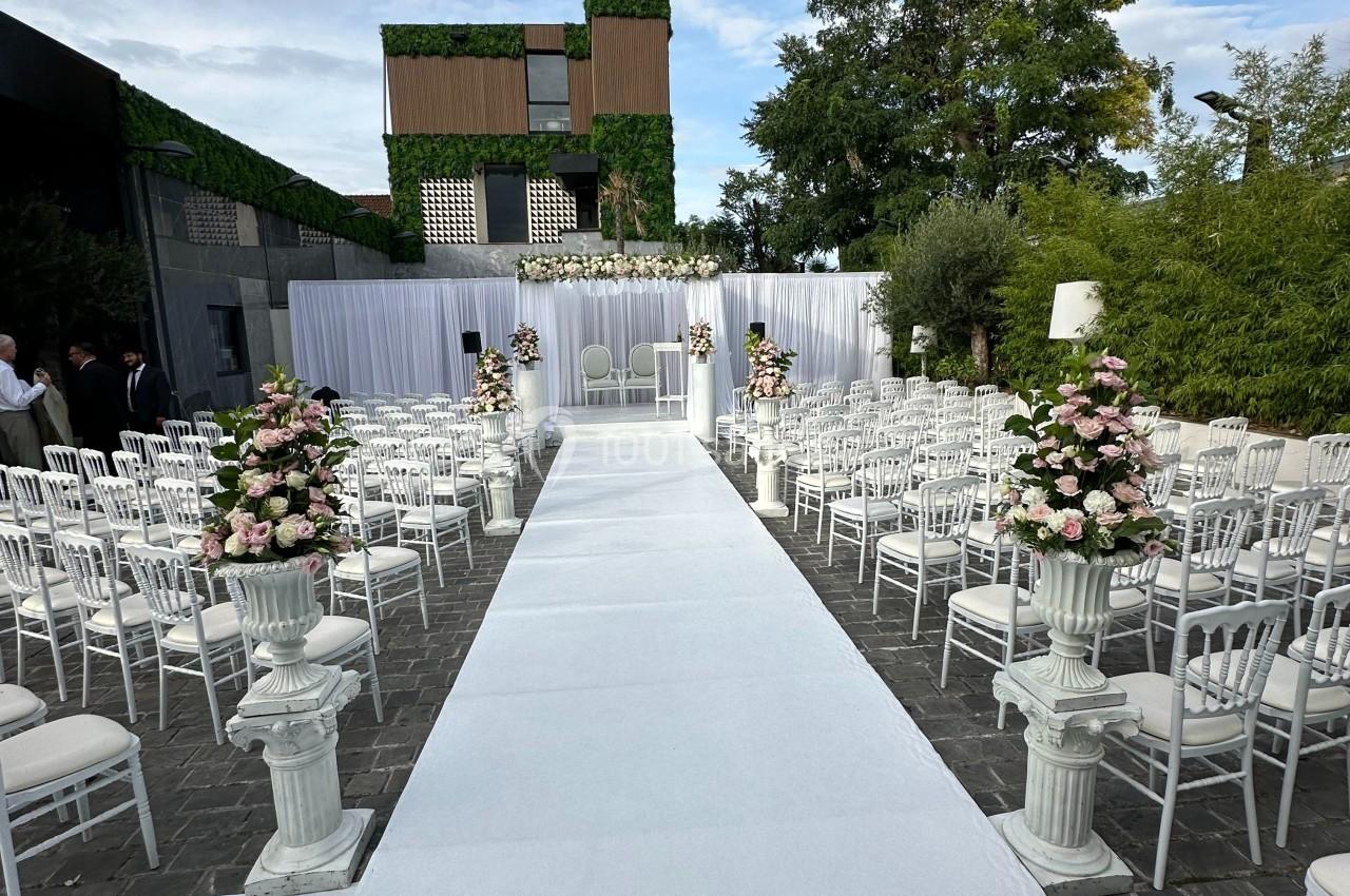 Allée centrale blanche bordée de chaises et de fleurs menant à un autel décoré pour une cérémonie en extérieur.