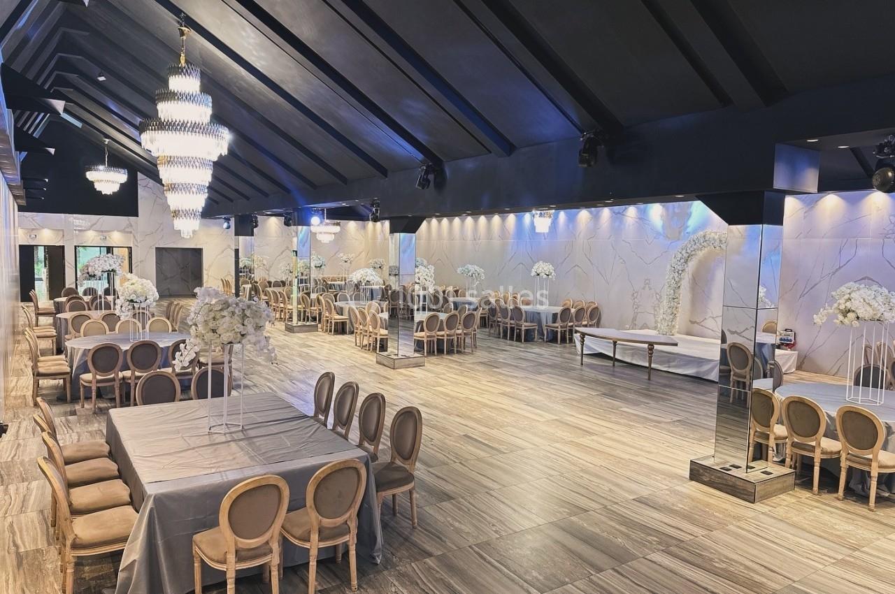 Salle de réception élégante avec tables dressées, chaises en bois clair, lustres suspendus et décorations florales blanches.