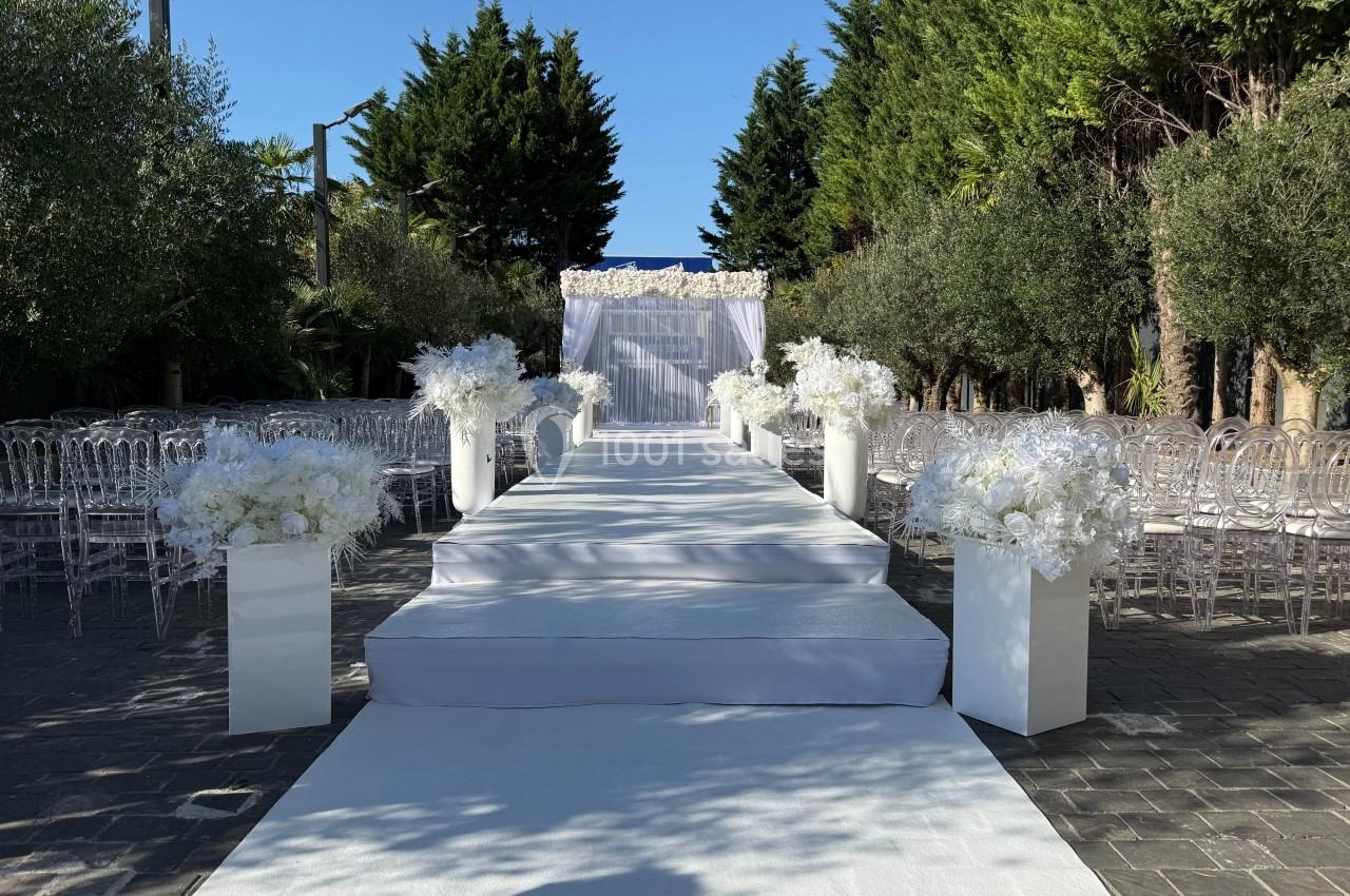 Allée de mariage extérieure décorée de fleurs blanches, avec une arche en toile blanche au bout, entourée de chaises…