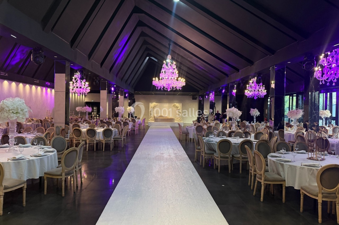 Salle de réception élégante avec tables rondes dressées, chandeliers suspendus et allée centrale blanche.