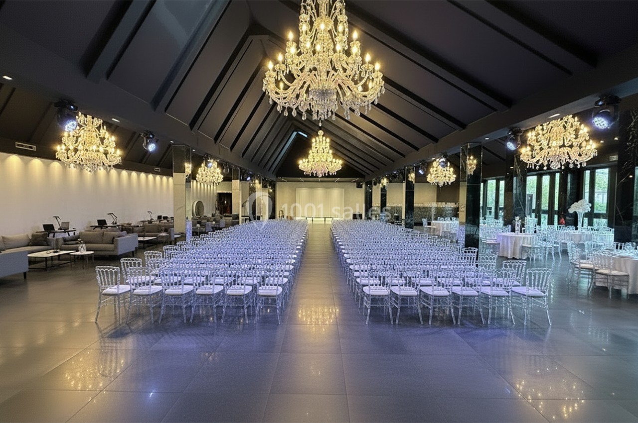 Salle de réception élégante avec rangées de chaises transparentes, lustres imposants et lumière naturelle en arrière-plan.