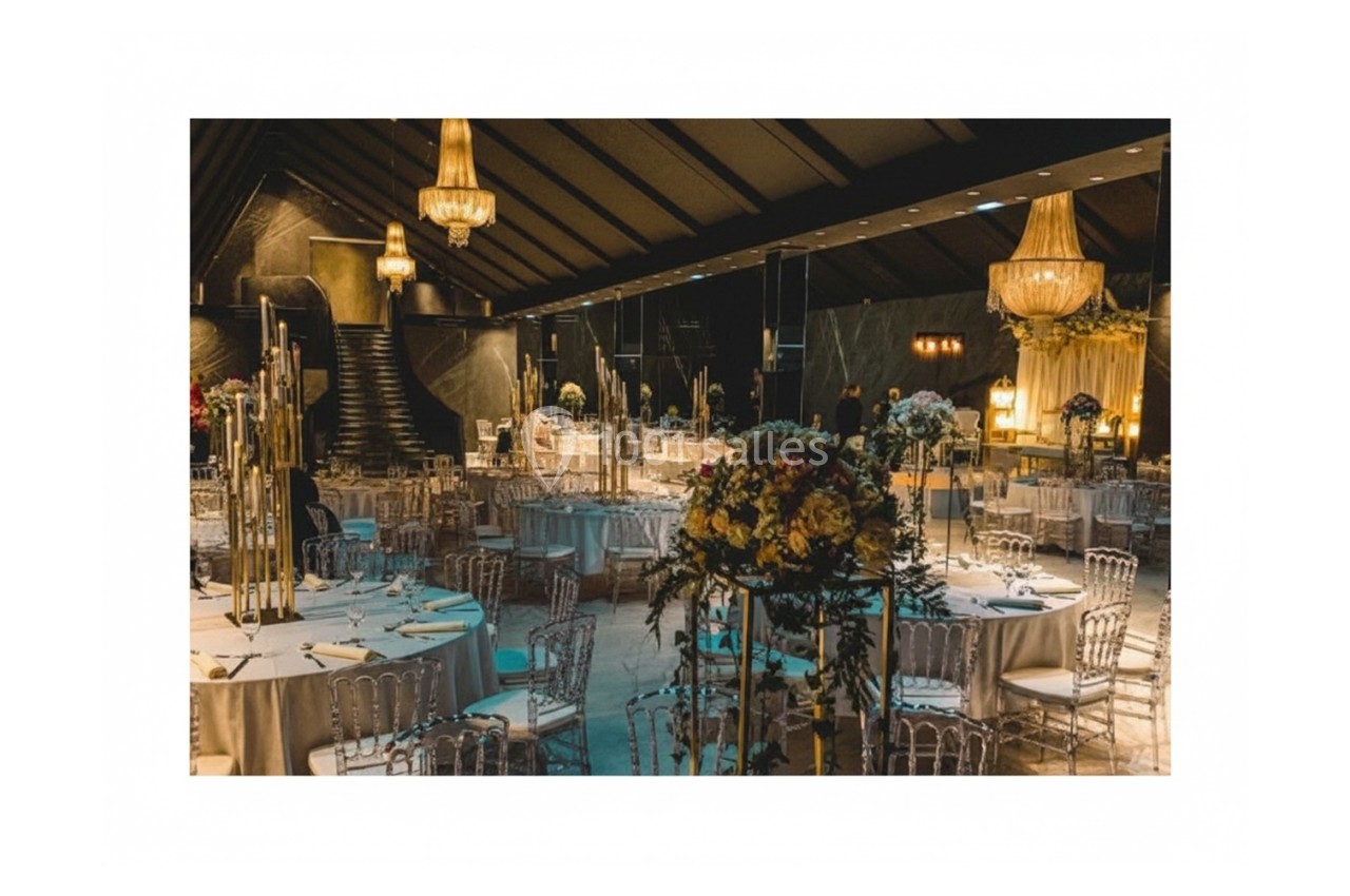 Salle de réception élégante avec tables dressées, chaises transparentes et lustres suspendus dans une ambiance tamisée.