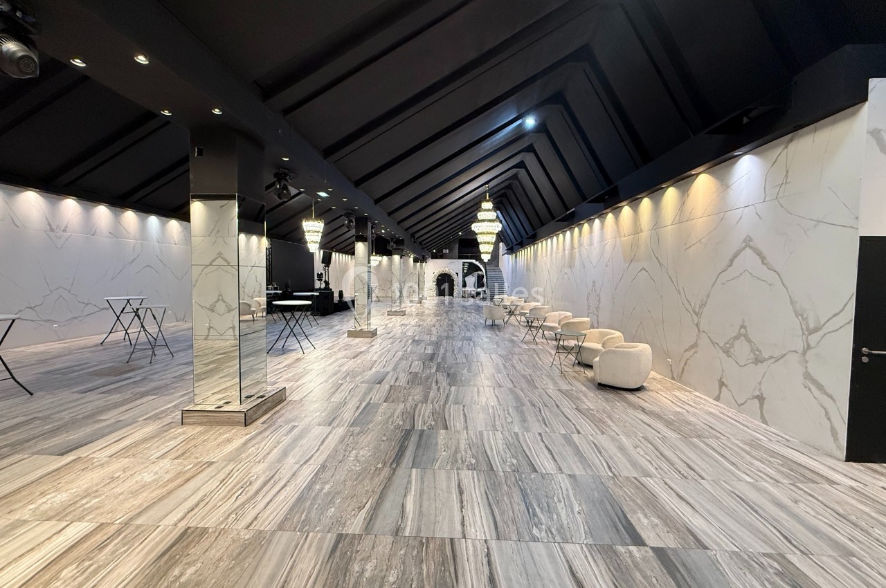 Salle spacieuse avec sol en marbre, murs clairs, plafonds sombres et lustres suspendus, aménagée avec tables et fauteuils.