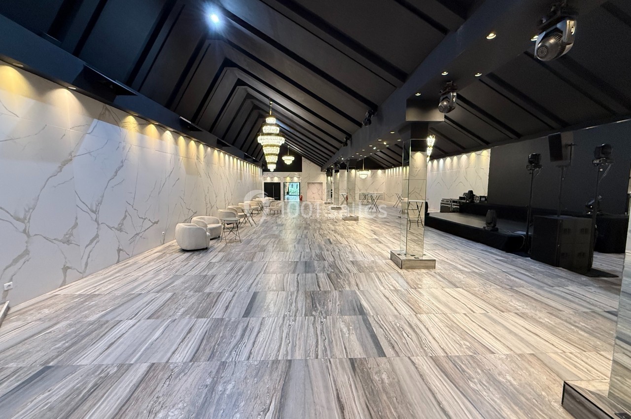 Salle spacieuse avec sol en marbre, murs clairs, lustres suspendus et scène équipée pour événements.