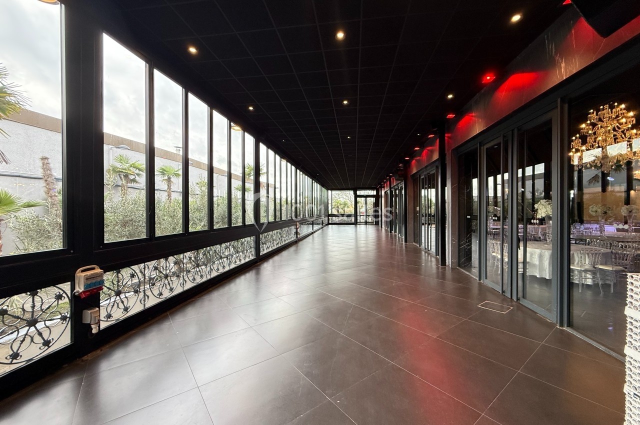 Couloir vitré avec sol en carrelage sombre, plafond noir et éclairage rouge, donnant sur une salle lumineuse.