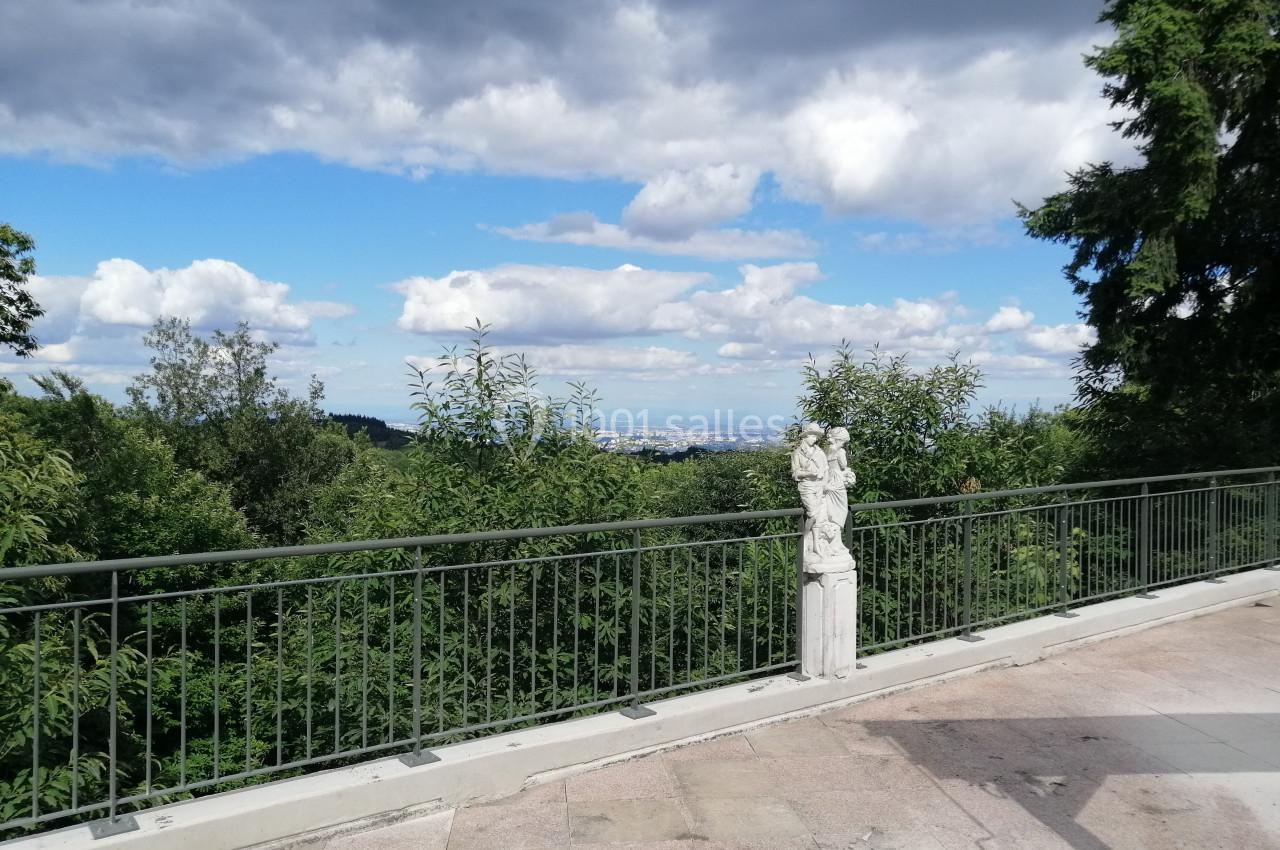 Vue d'une terrasse avec une statue, une balustrade, des arbres et un ciel partiellement nuageux en arrière-plan.