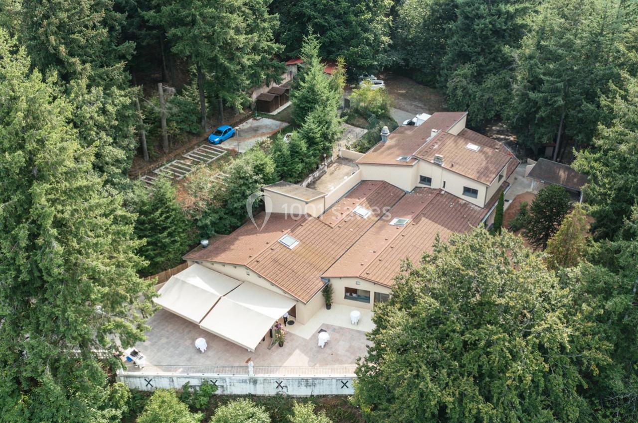 Vue aérienne d'une grande maison beige avec toit en tuiles, entourée de forêt et d'une terrasse aménagée.