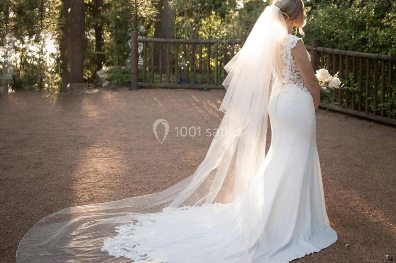 Une mariée en robe blanche avec une longue traîne et un voile se tient dans un jardin ensoleillé près d'une barrière en bois.