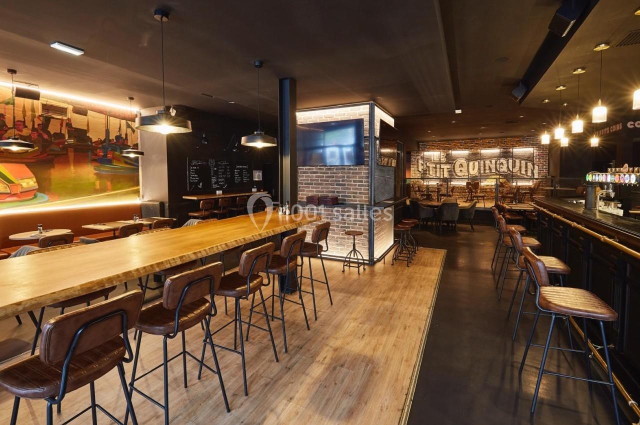 Intérieur d'un bar-restaurant moderne avec comptoir, tables en bois, chaises hautes et éclairage chaleureux.