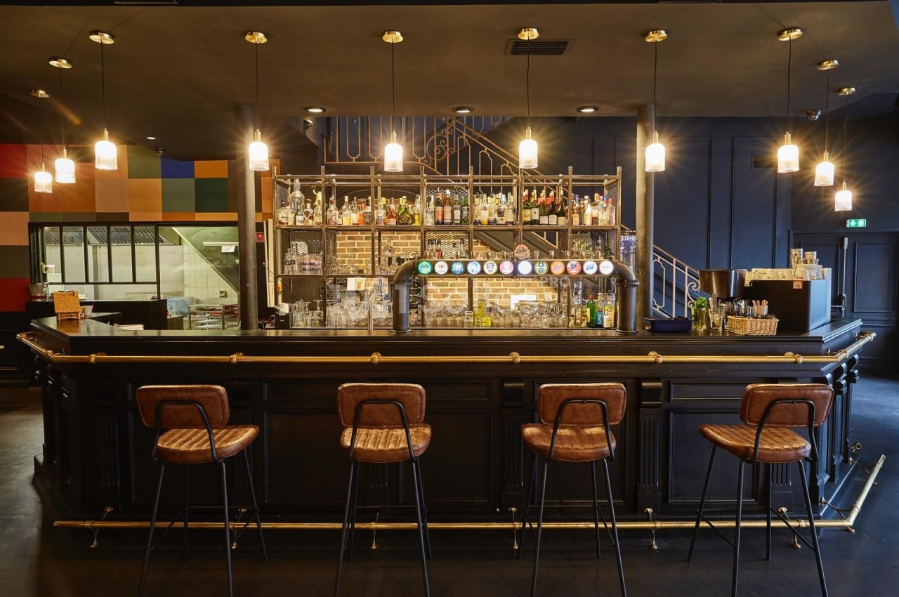 Bar avec comptoir en bois sombre, tabourets hauts, étagères de bouteilles et lumières suspendues dans une ambiance…