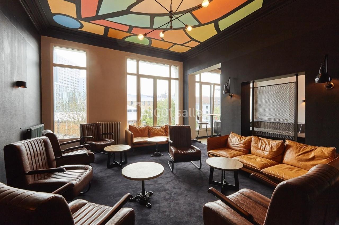 Salle de réunion avec fauteuils en cuir, tables basses, plafond coloré et grandes fenêtres laissant entrer la lumière…