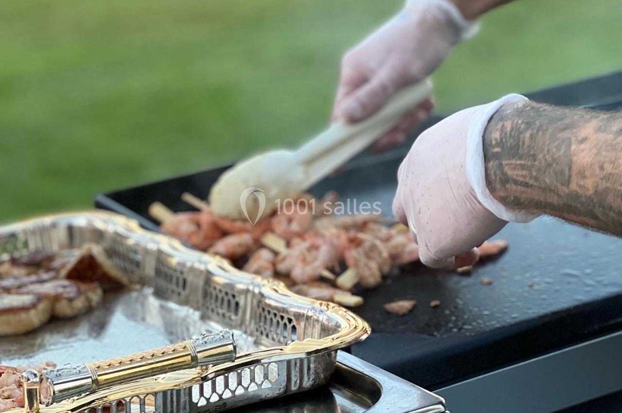 Personne cuisinant des brochettes de crevettes sur une plancha avec un plateau de viande à proximité.