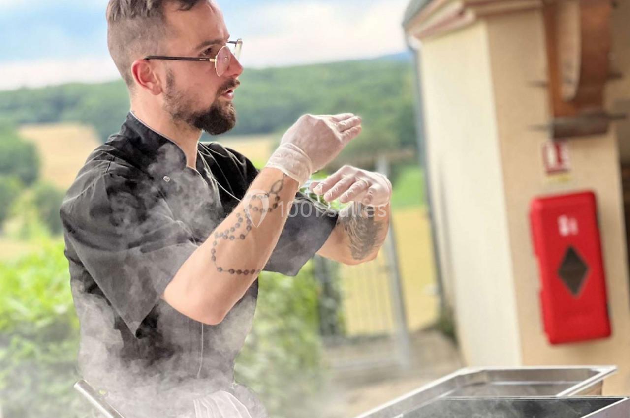 Un chef cuisinier en tenue noire prépare un plat en extérieur, entouré de vapeur, avec un paysage rural en arrière-plan.