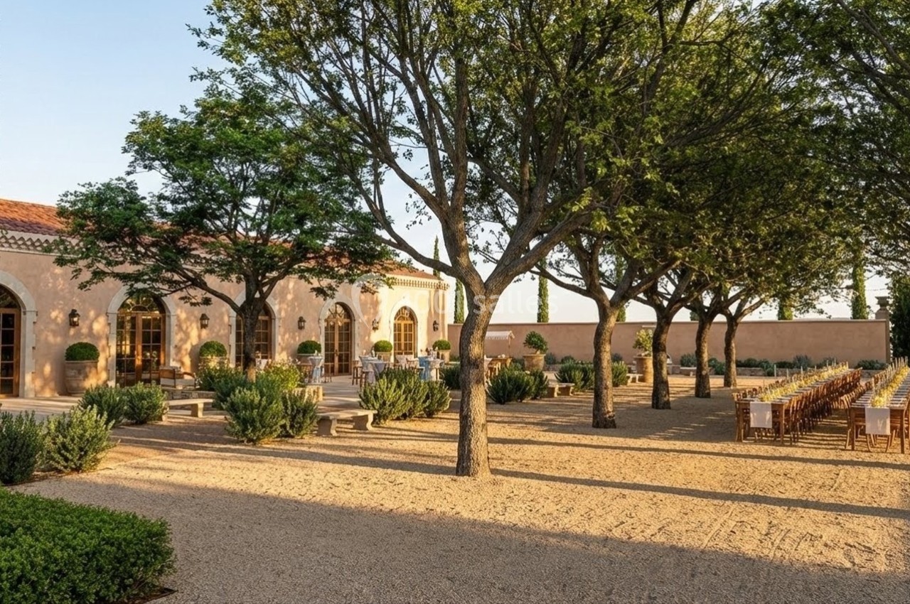Allée bordée d'arbres menant à une terrasse avec tables dressées et bâtiment en pierre aux grandes fenêtres.