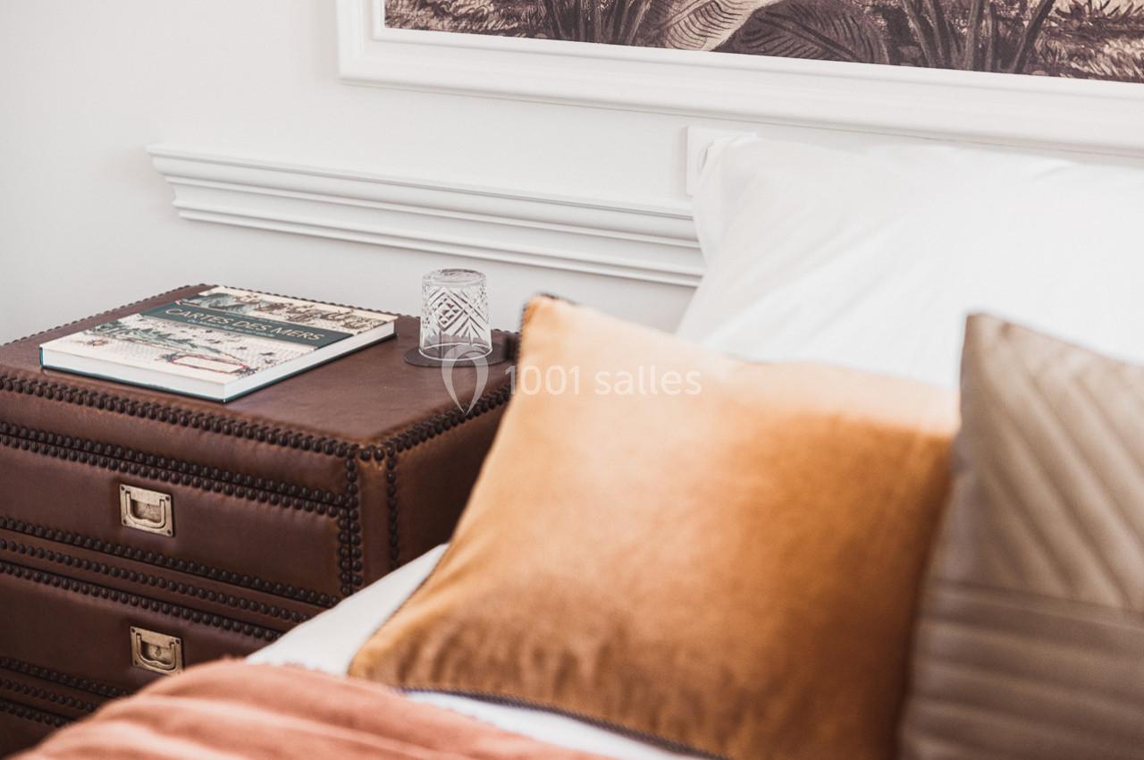 Table de chevet en cuir avec un livre et un verre, à côté d'un lit aux coussins beige et orange dans une chambre lumineuse.