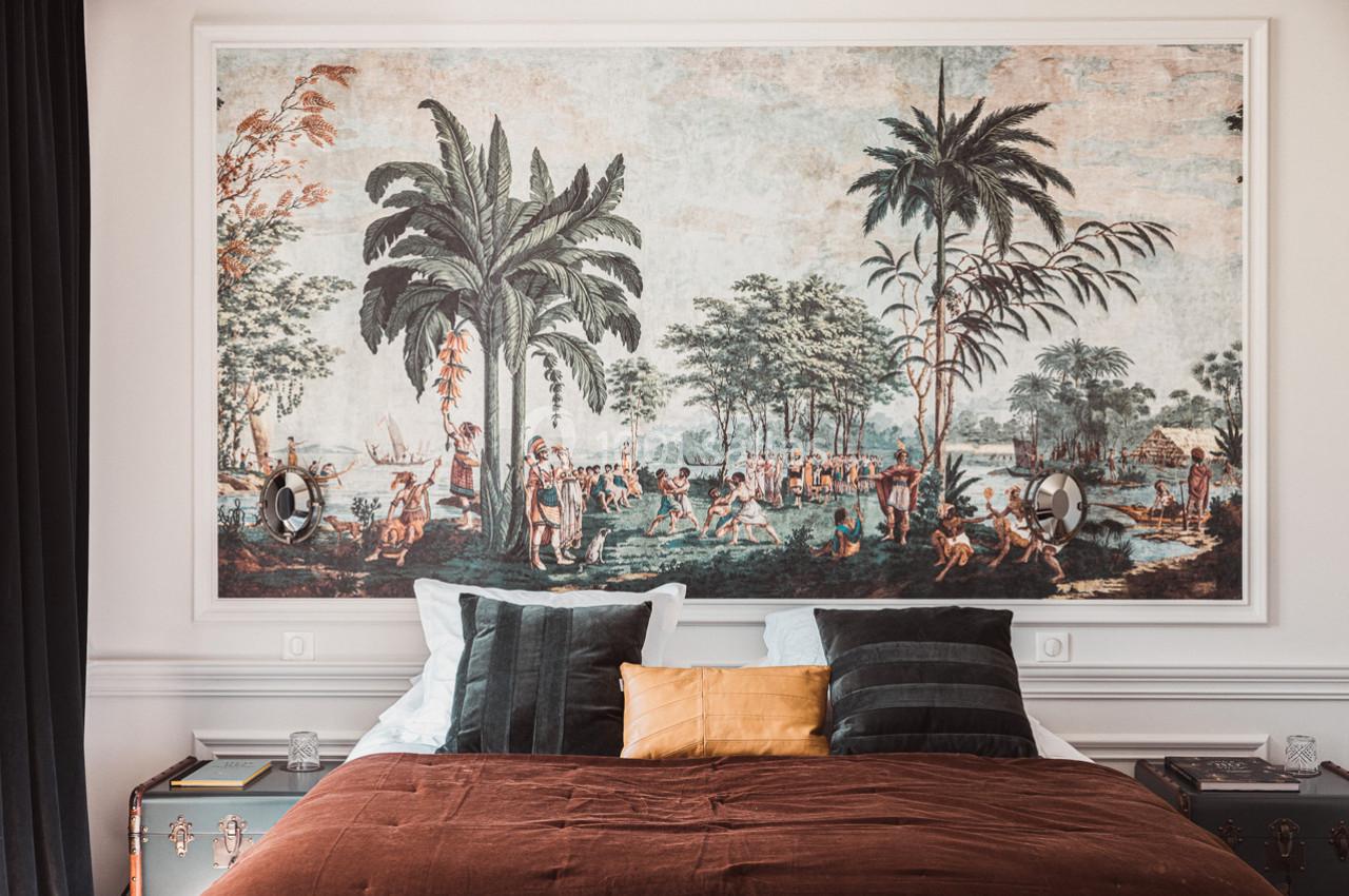 Lit avec coussins devant une grande fresque murale représentant une scène tropicale avec palmiers et personnages.