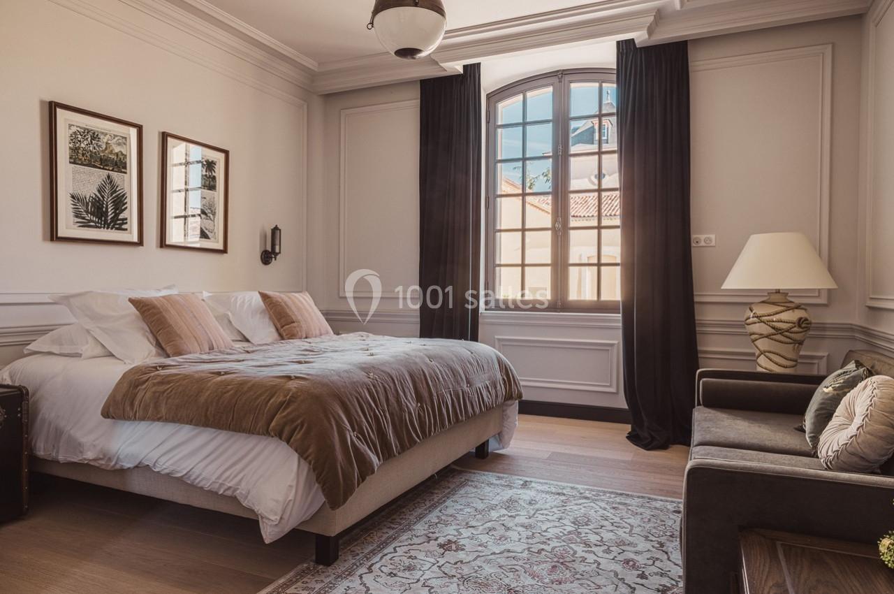 Chambre lumineuse avec lit double, rideaux sombres, fauteuil, lampe et décorations murales végétales.