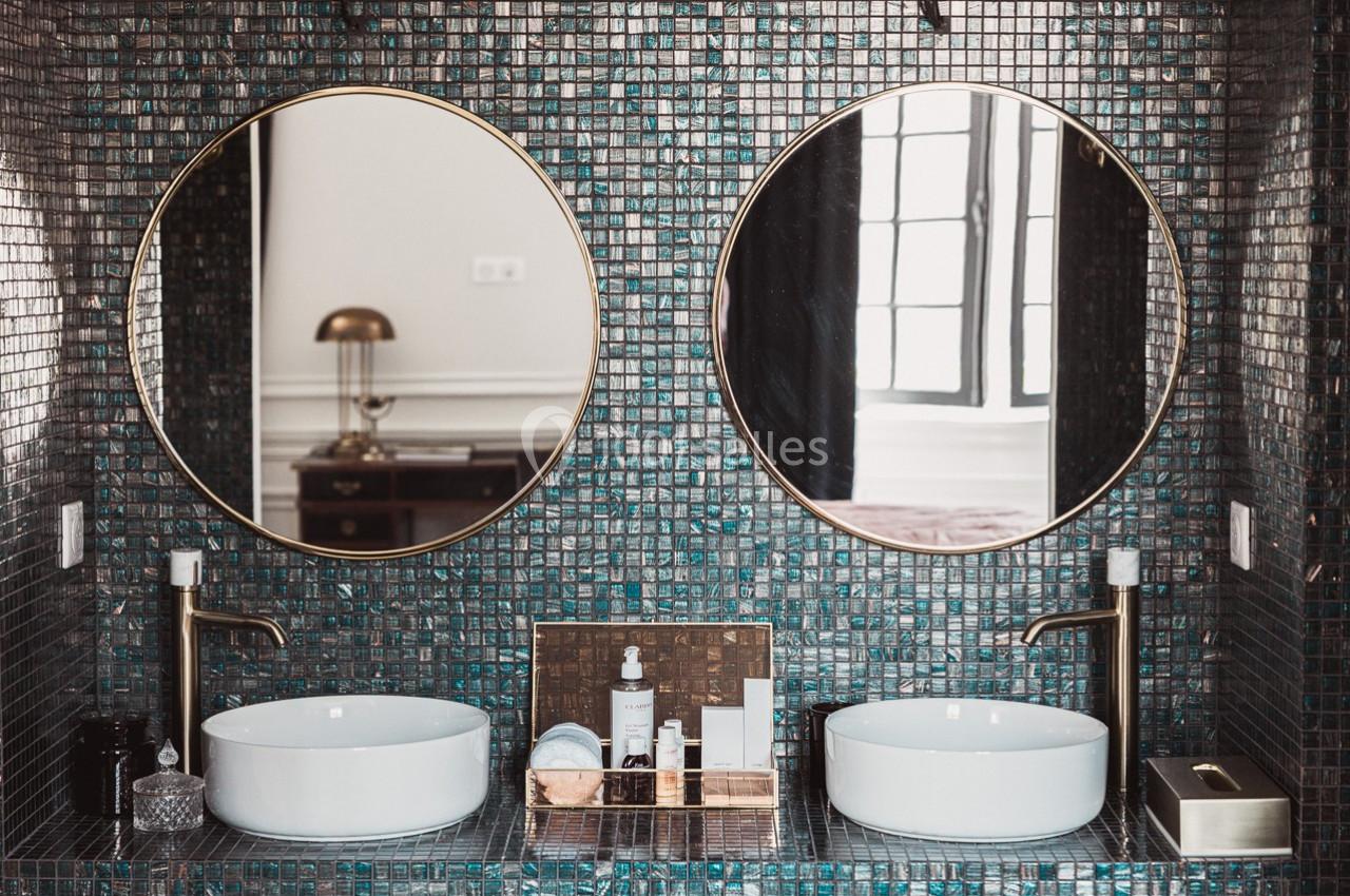 Deux lavabos blancs avec robinets dorés devant des miroirs ronds, sur un mur de carreaux sombres brillants.