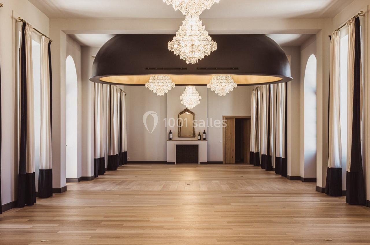 Salle spacieuse avec parquet clair, hauts plafonds, lustres en cristal et grandes fenêtres encadrées de rideaux.