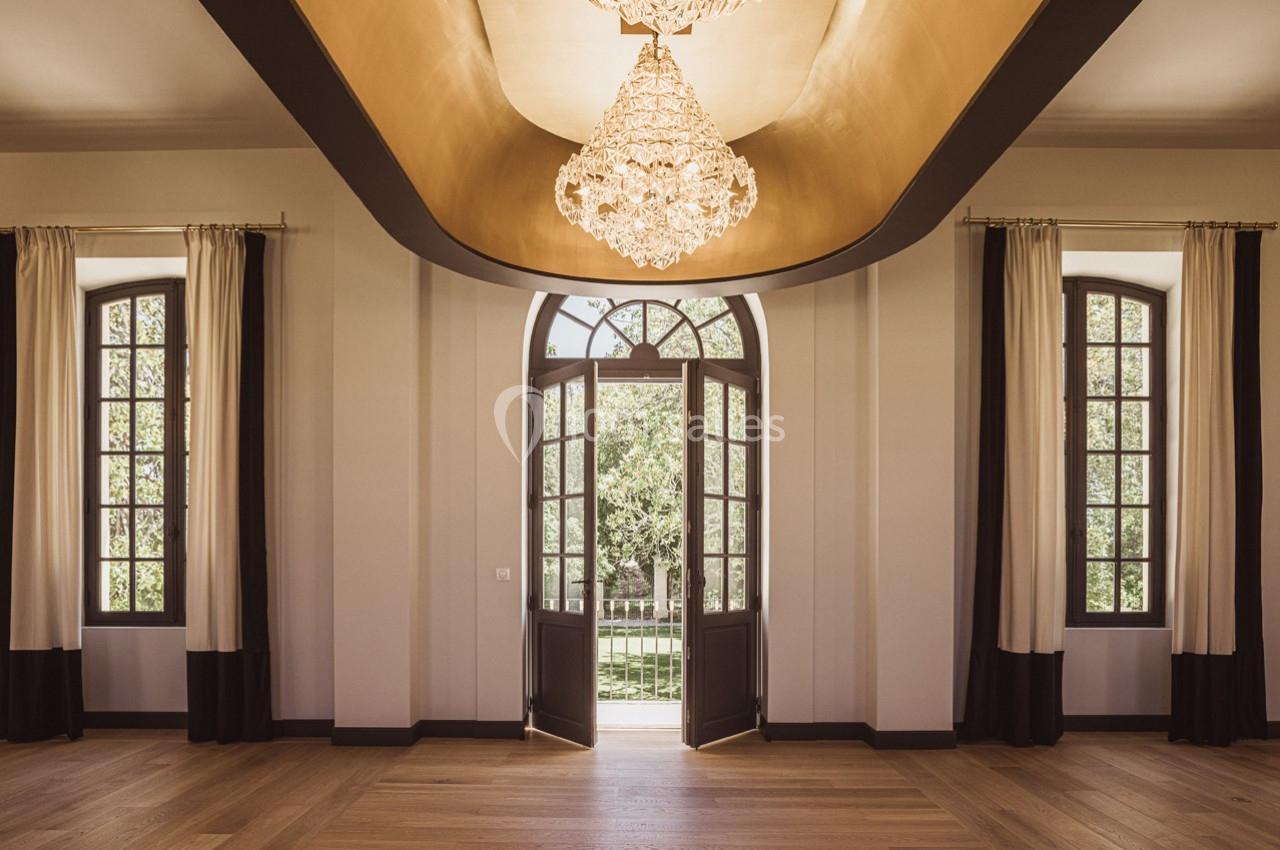 Vue intérieure d'une pièce lumineuse avec un lustre central, de grandes fenêtres et une porte ouverte sur un jardin.