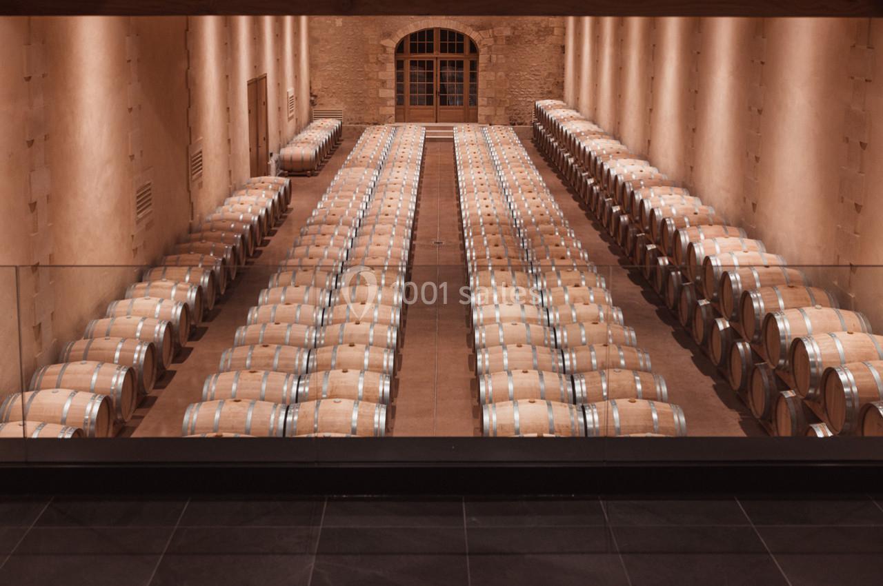 Rangées alignées de fûts en bois dans une cave voûtée éclairée, avec un mur en pierre au fond.