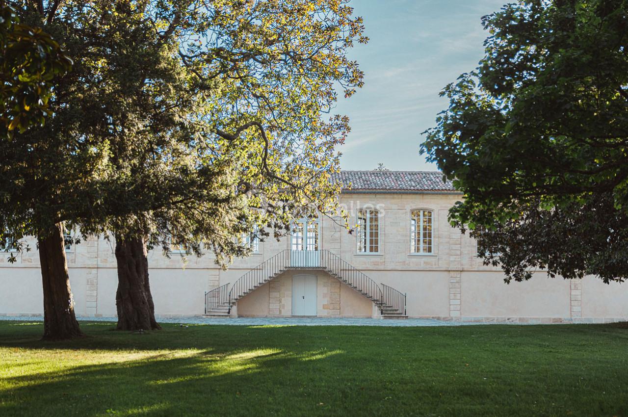 Façade d'un bâtiment en pierre avec escalier central, entouré d'arbres et d'une pelouse verdoyante.