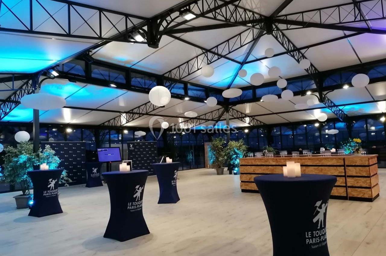 Salle lumineuse avec un plafond en structure métallique, décorée de lanternes suspendues et de tables hautes drapées.