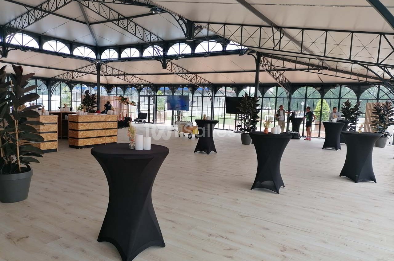 Salle lumineuse avec parquet clair, tables hautes noires, plantes en pot et grandes baies vitrées.