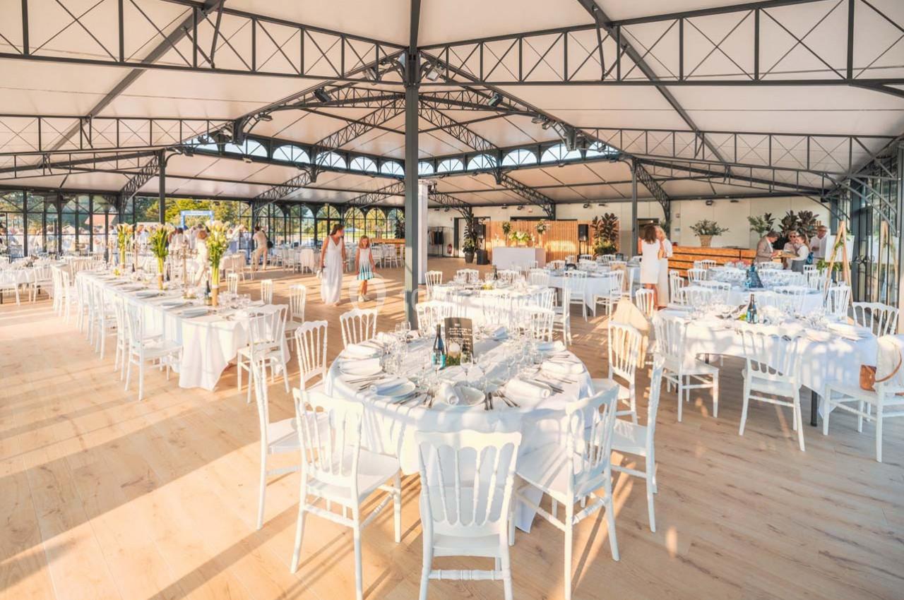 Salle de réception lumineuse avec tables rondes dressées, chaises blanches et structure métallique apparente.