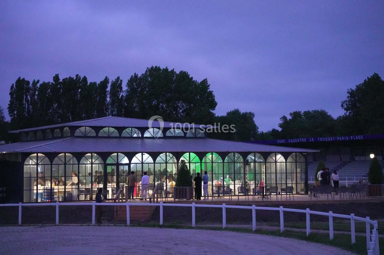 Bâtiment vitré éclairé en soirée, entouré d'arbres et d'une clôture blanche, avec quelques personnes à l'extérieur.