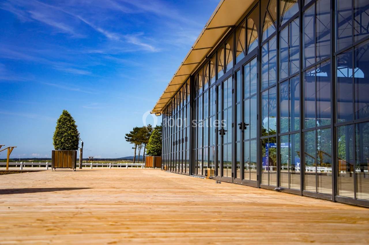 Grande terrasse en bois bordant un bâtiment vitré sous un ciel bleu dégagé, avec quelques arbres en arrière-plan.