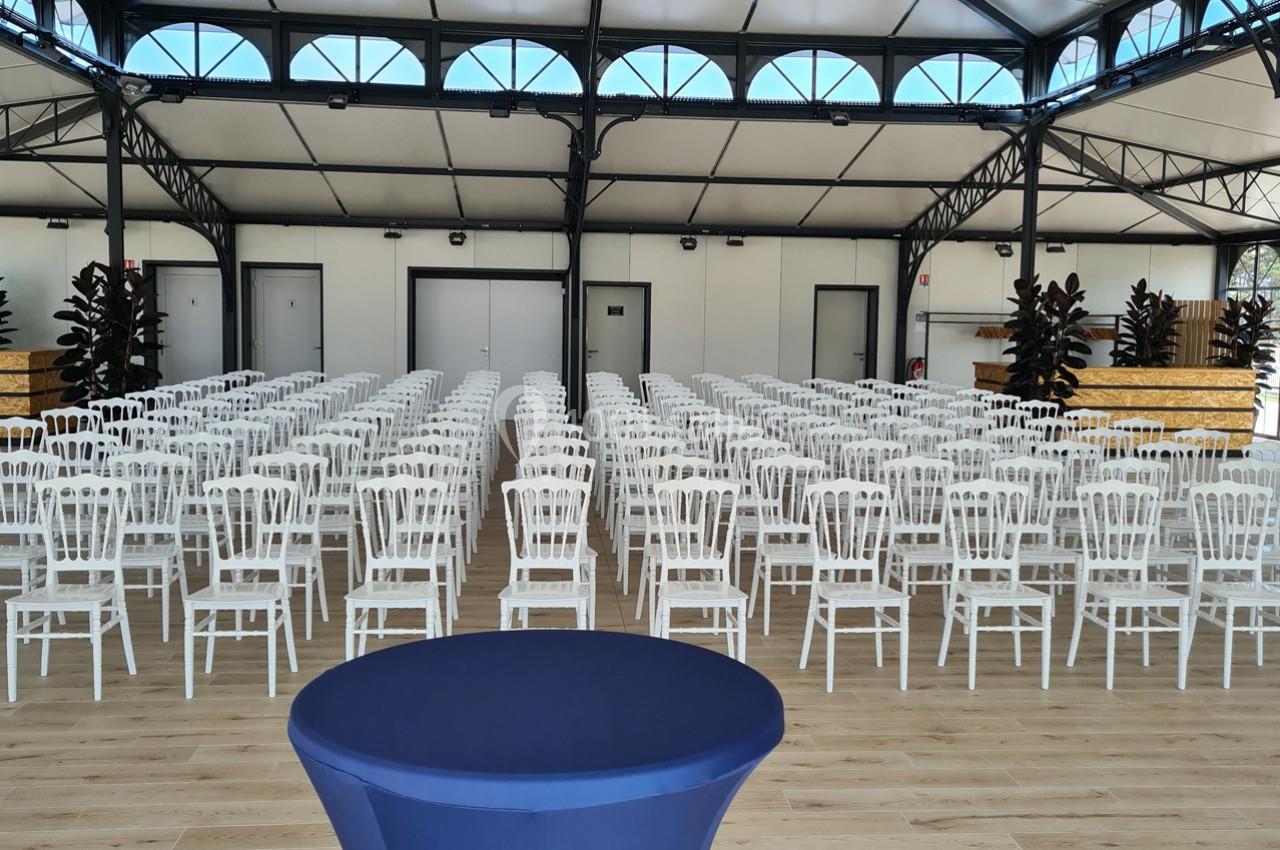 Salle aménagée avec des rangées de chaises blanches alignées devant une table recouverte d'une nappe bleue.