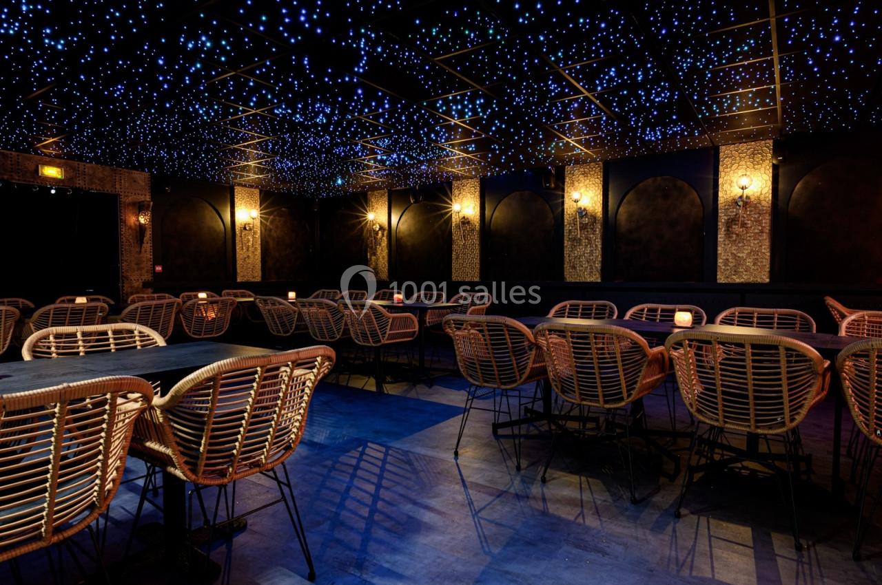 Salle sombre avec des tables et des chaises en rotin, éclairée par un plafond étoilé et des appliques murales.