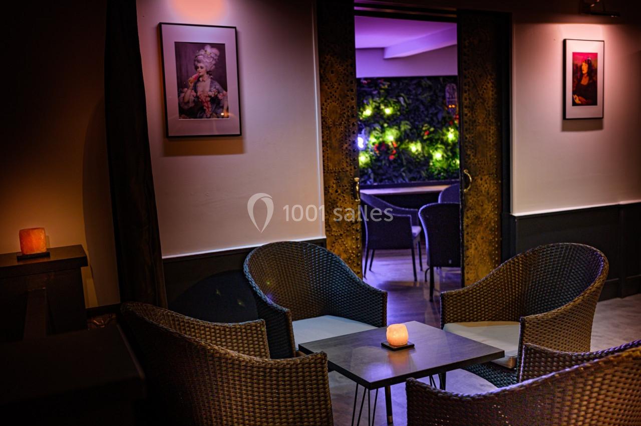 Espace intérieur avec fauteuils en rotin autour d'une table, vue sur une terrasse éclairée et décorée de plantes.