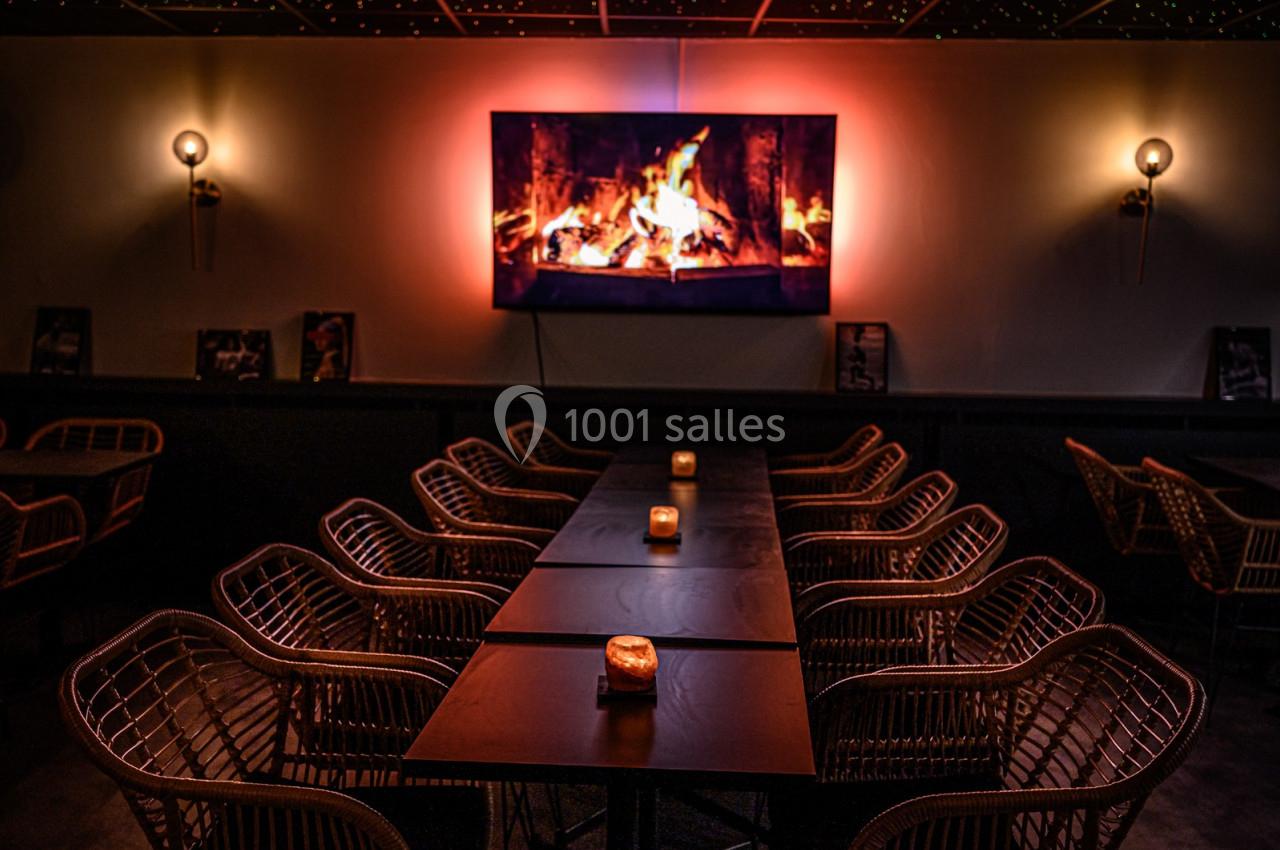 Salle sombre avec des tables alignées, des chaises en rotin et un écran affichant une cheminée enflammée.